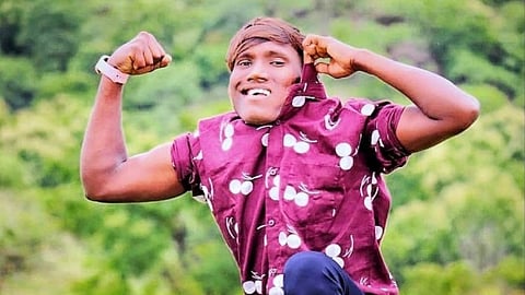 Suraj Chavan crouches on grass, flexing muscles and smiling confidently. They wear a maroon shirt with white patterns. Green trees fill the background.