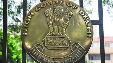 Brass emblem of the Delhi High Court on a gate, featuring the Ashoka Lion capital, with greenery and a building in the background, conveying authority.