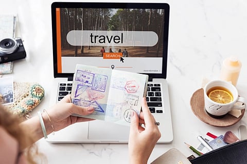 A desk- a laptop with the screen displaying a travel location with travel written as the caption, camera, coffee mug and a winter hat on the desk, also a pair of hands (most likely a woman's) holding a passport