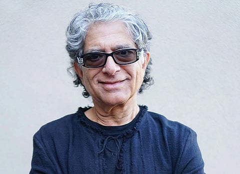 Image of New Age guru Deepak Chopra. In the image Chopra is with gray hair and glasses smiles, wearing a dark shirt with textured details, against a plain light background.