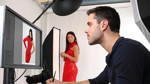 A photographer edits a photo on a computer in a studio. A model in a red dress stands in the background. The scene conveys focus and creativity.