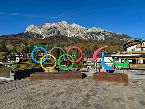 Olympic and Paralympic symbols stand on a terrace, with picturesque mountains and a clear blue sky in the background, conveying a sense of awe.