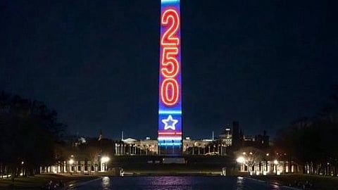 The Washington Monument illuminated at night with red and blue lights displaying "250" and stars, reflecting in a calm pool. Celebratory and patriotic mood.