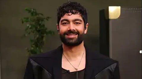 32nd Avenue Founder Dhruv Dutt Sharma with curly hair and a beard, wearing a black outfit and necklace. A plant and a modern decorative element are in the dimly lit background.