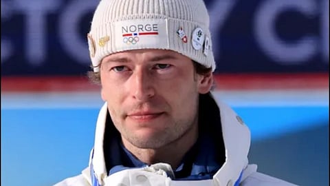 Norwegian biathlete Sturla Holm Lægreid in a white jacket and beanie with Norway logo, stands solemnly with a medal lanyard. Vibrant background suggests a winter sports setting.