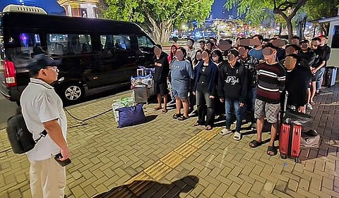 A group of people stand in an orderly row beside a parked van at night, facing a person in a white shirt. Luggage and bags are beside the van. The setting appears calm and organized.