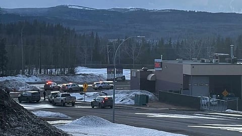 Police vehicles with flashing lights are parked near a snow-covered roadside by a building. Mountains and forests are visible in the background. The scene appears tense.