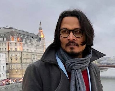 Image of youtuber Ajeet Bharti glasses standing near a river, wearing a scarf and coat. In the background are ornate buildings under an overcast sky.