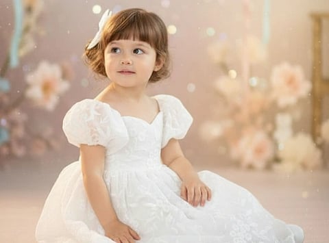 A young child in a white dress with puffed sleeves sits charmingly. Soft focus background with pastel flowers and gentle bokeh, creating a dreamy, whimsical feel.