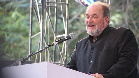 Author William Dalrymple in a dark coat speaks at a podium outdoors, under colorful stage lights. He appears confident and engaged, with a backdrop of trees and metal scaffolding.