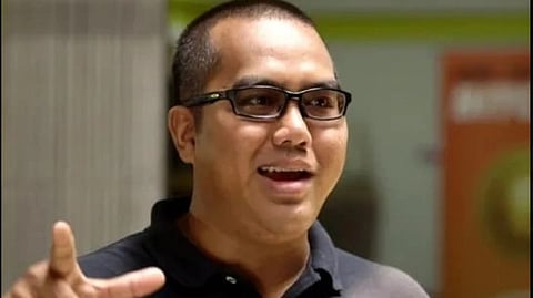 Malaysian entrepreneur Arsyan Ismail with glasses and a buzz cut is smiling and gesturing with one hand. He wears a dark polo shirt. The background is blurred with warm tones.