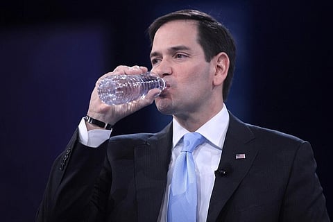 Image of US delegation led by Secretary of State Marco Rubio in a suit and light blue tie drinks from a water bottle on stage against a dark background