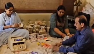 Three people sit on a bed surrounded by stacks of currency notes. A counting machine is nearby, indicating they are engaged in handling money. The scene feels tense and focused.