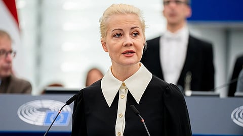 Yulia Navalnaya in formal attire speaks at a microphone in a conference setting, with a backdrop of blurred figures and a logo. She appears focused and confident.