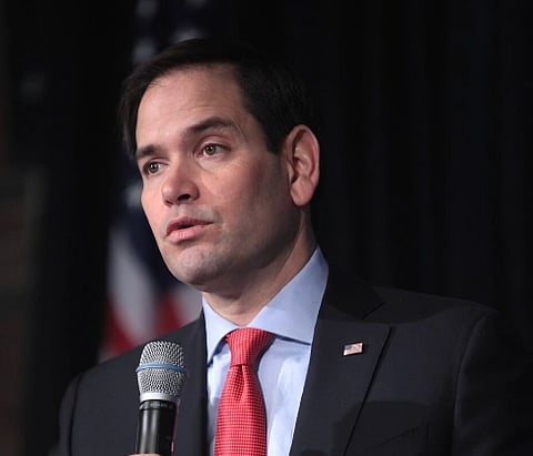 Image of Marco Rubio speaking at an event in Manchester, New Hampshire. He is wearing a suit and red tie speaking into a microphone, with an American flag blurred in the background.
