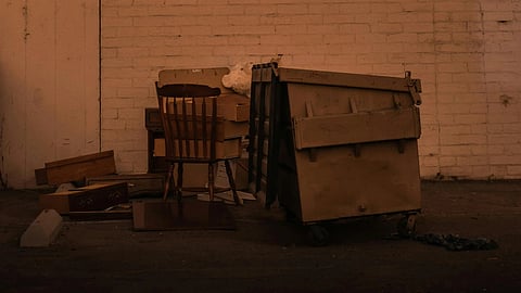 Orange-tinted sunset illuminates an urban alley. A wooden chair and dumpster are against a brick wall, casting deep shadows, evoking a sense of solitude.
