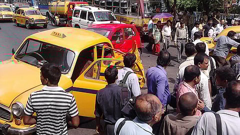 A crowded street scene with a damaged yellow taxi involved in an accident. People gather around, appearing concerned, as other vehicles and buses are nearby.