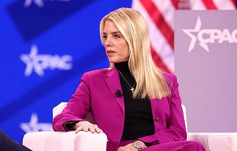 Attorney General Pam Bondi in a vibrant magenta suit sits on a stage with a thoughtful expression. Background features a CPAC logo and American flag