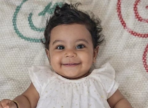 Image of 10-month-old baby girl, Alin Sherin Abraham. She has curly hair smiles widely, lying on a textured blanket with green and red swirl patterns.