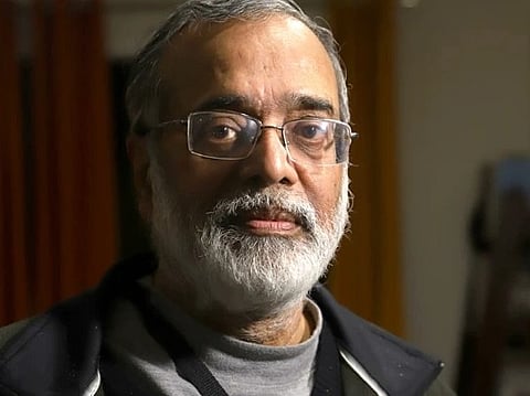 Image of Journalist Prabir Purkayastha with glasses and a beard, wearing a jacket over a gray shirt. He stands indoors with a neutral expression