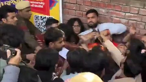 Crowded scene with people against a brick wall, some showing distress. Police officers, one in uniform and cap, manage the crowd in a tense atmosphere.