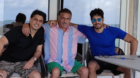 Actor Sidharth Malhotra with his father and brother sitting together, smiling, with arms around each other. They are at an outdoor table by the sea, reflecting a relaxed and joyful mood.