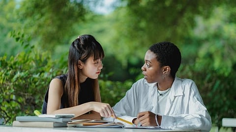 Two people sit at an outdoor table surrounded by greenery, engaged in a focused discussion over open books and notes, conveying collaboration and study.