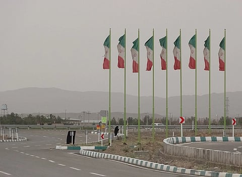 Image of National flags in Iran, Qom city. A row of Iranian flags on tall poles lines a curving road under a cloudy sky. Mountains are visible in the distance, creating a serene atmosphere.