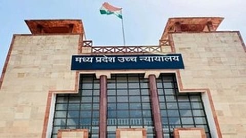 A large stone building with a modern facade features tall columns and a sign in Hindi. An Indian flag waves on the roof under a clear sky.