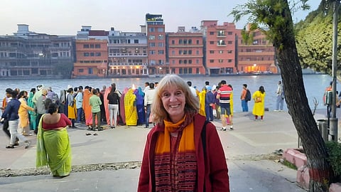 Maria Wirth smiling in front of a riverbank with a crowd gathered, wearing colorful attire. Behind her, vibrant buildings line the water's edge, creating a lively atmosphere.