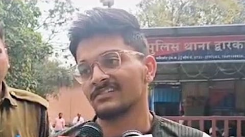 Accused Piyush Dhamnotiya with glasses speaks into microphones from various media outlets outside a police station. An officer stands beside him, trees and the station sign in the background.