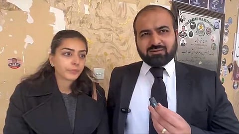 Human Rights Lawyers of Pakistan Imaan Zainab Mazari-Hazir and Hadi Ali Chattha in formal wear stand against a peeling, worn wall with various stickers and posters. The man holds a microphone, and both appear serious.