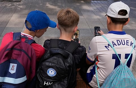 Three boys sit closely on a bench, absorbed in their smartphones. Each wears a sporty backpack, with jerseys and caps adding a casual vibe.