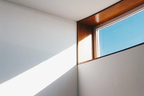 Sunlight streams through a wooden-framed window, casting a sharp diagonal shadow on a white wall, creating a calm and minimalistic atmosphere.