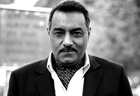 A man in a suit and polka dot scarf stands outdoors, looking confidently at the camera. The image is in black and white, with a soft-focus background.