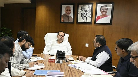 A group of eight individuals in a formal meeting around a wooden table, with documents and coffee cups. Portraits on the wall suggest an official setting.