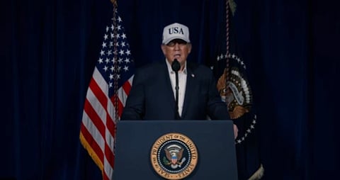 President Donald Trump in podium with US Army Sign behind him are two flags of America and American Army and he is wearng a USA cap