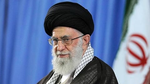 In the image Ayatollah Ali Khamenei is shown looking at the camera behind him is a blue background and the flag of Iran