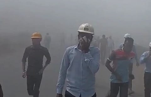 Image of workers running with their mouth covered with cloth. A dense fog is seen behind them. the people seem to be running in panic.