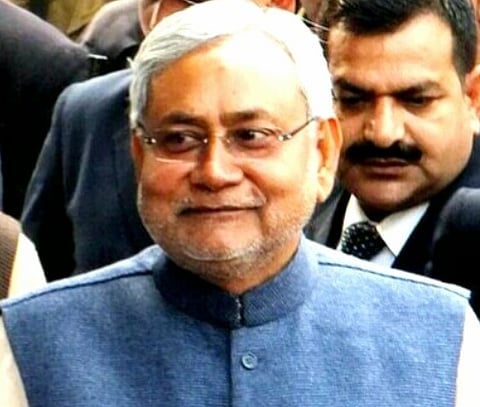 Image of Bihar cm Nitish Kumar wearing a blue jacket, smiles slightly. He is surrounded by people in suits, creating a formal atmosphere.