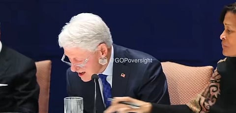 Image of former US president Bill Clinton grinning over a photograph during his deposition. His lawyer sitting on his left seems to be taking that photograph away from him.