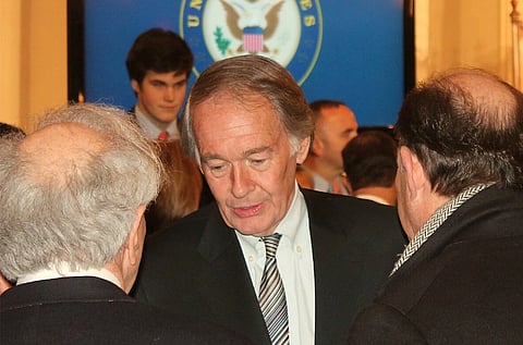 Image of Sen. Markey talking to other people. he is in a room filled with people. He speaks with two older men in a crowded room. The U.S. emblem is visible on a screen in the background, indicating a formal event.