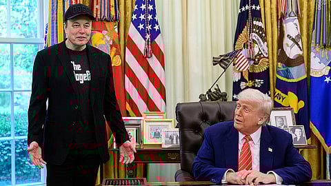 Elon Musk in all black standing next Donald Trump as he is sitting in the Oval Office