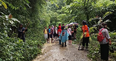 A group of people is standing on a muddy path in a lush, green forest. Some hold umbrellas. The atmosphere is adventurous and lively.