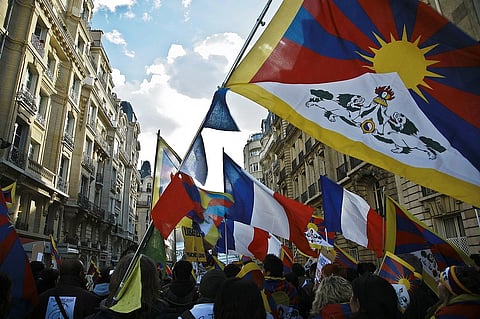 Protest scene with Tibetan flags and French flags waving, set between historic Parisian buildings. The atmosphere conveys unity and activism.