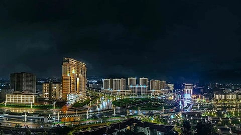 Nighttime cityscape with illuminated high-rise buildings, streets, and vibrant lights reflecting on wet surfaces. The scene conveys an energetic urban vibe.