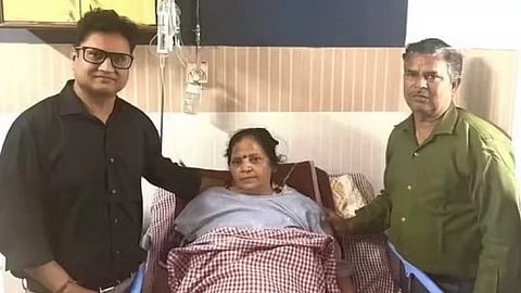 In the image a hospital is shown where a woman is lying in a ventilator and two men are standing on either side of her everyone is looking at the camera