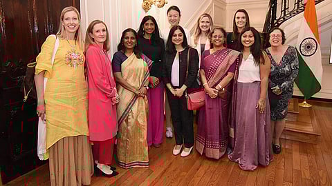 In the image Indian Americans are shown wearing colourful clothes looking at camera with behind them is an Indian Flag
