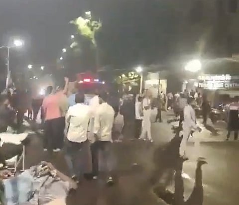 A nighttime street with a crowd gathered. People are standing and some are gesturing with raised hands. Streetlights and vehicles are visible.