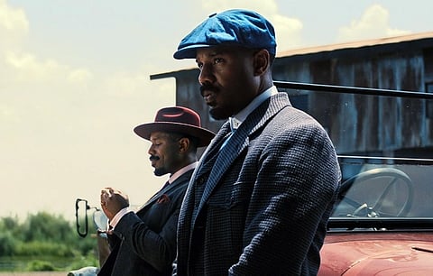 Two men ( Hollywood actor Michael B. Jordan) in vintage attire stand by an antique car. The man in the foreground wears a blue cap; both have serious expressions in a rustic setting.